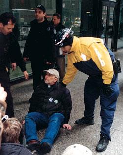 Greg Dell seated on the pavement with others while a police officer bends over him, encouraging him to get up and move.