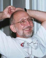 Greg Dell in white T-shirt leaning back with his hands behind his head and smiling.