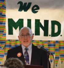 Greg Dell in front of a "We M.I.N.D." banner.