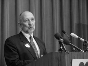 Black and white photo of Greg Dell standing a a podium with microphones.